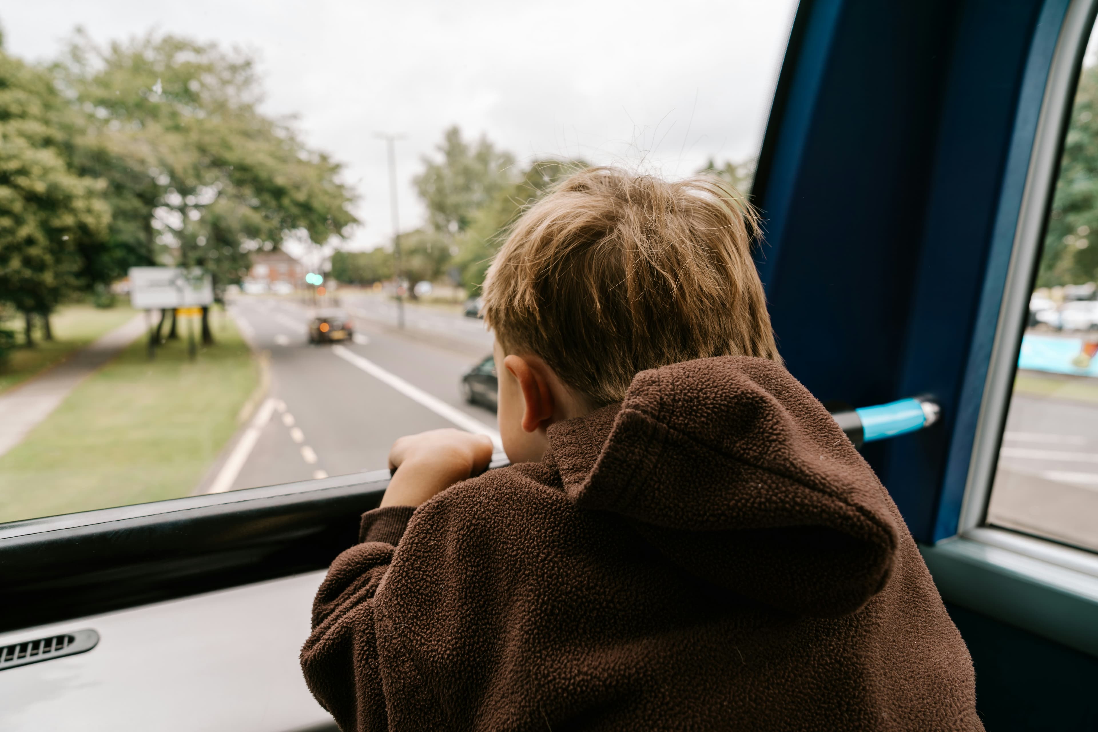 Child in a brown hoodie looks out the window of a moving bus at a tree-lined street.