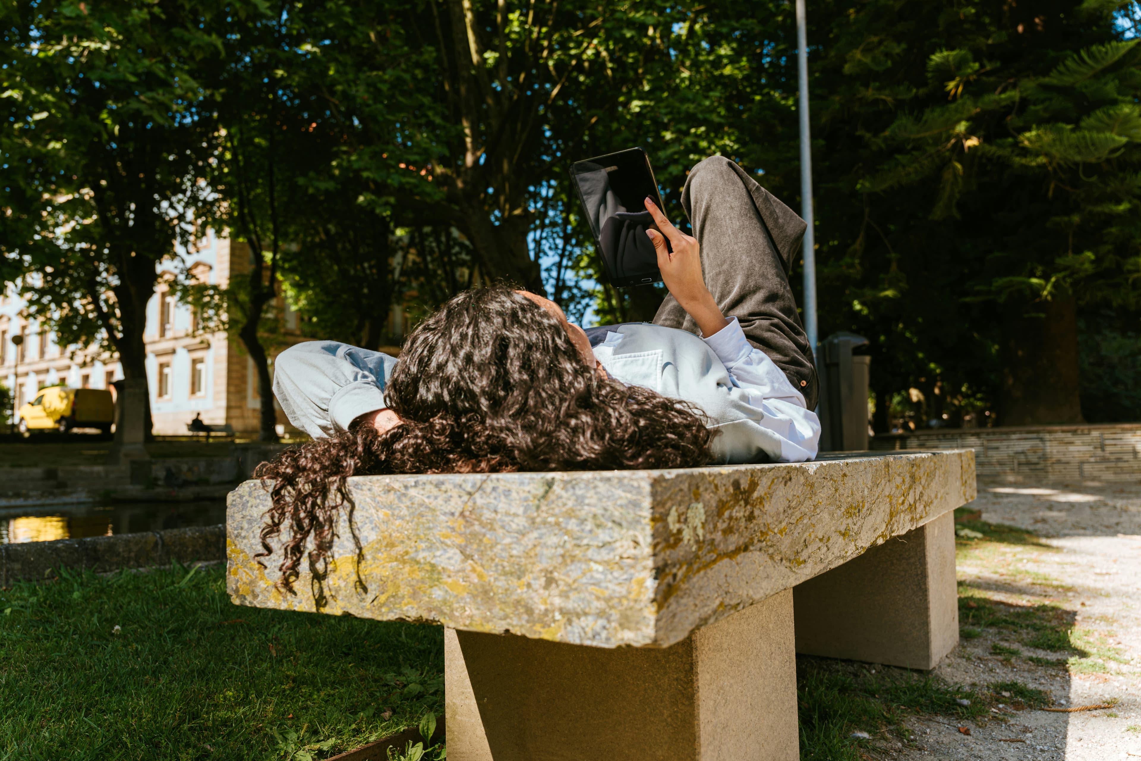 Person with curly hair lying on a stone bench in a park, looking at a smartphone, surrounded by trees and sunlight.