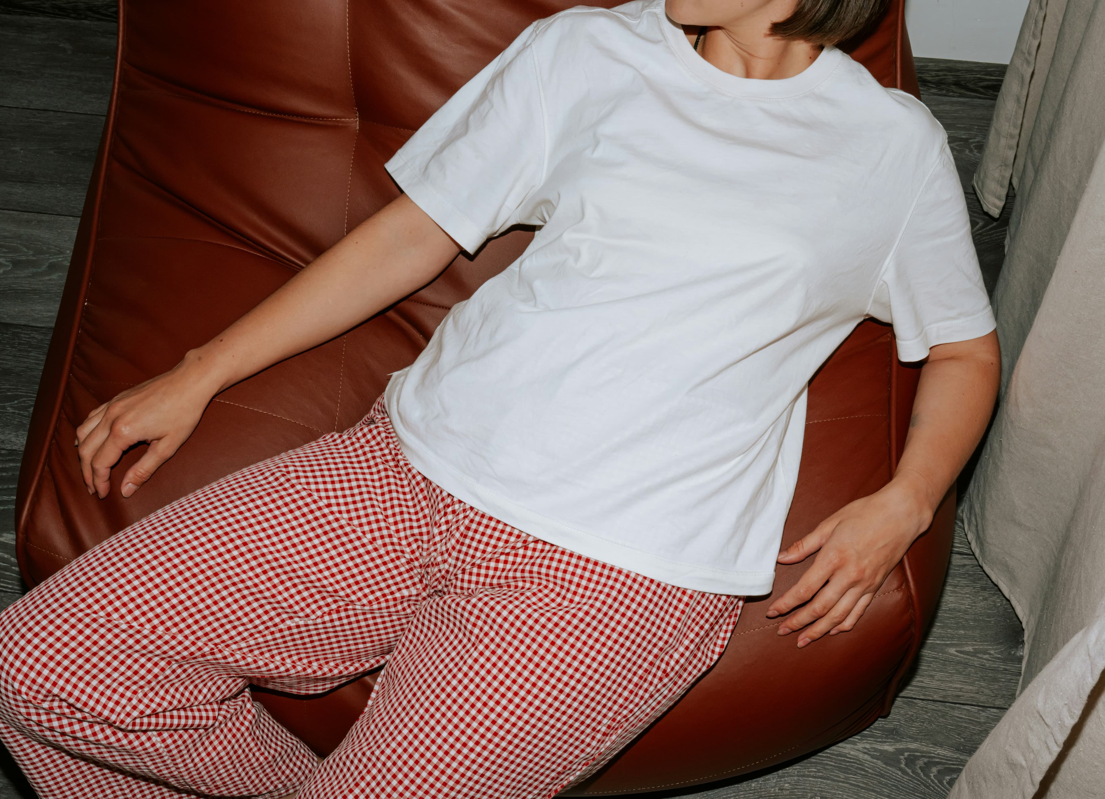 Person in a white t-shirt and red checkered pants lounging on a brown chair, with a relaxed posture.